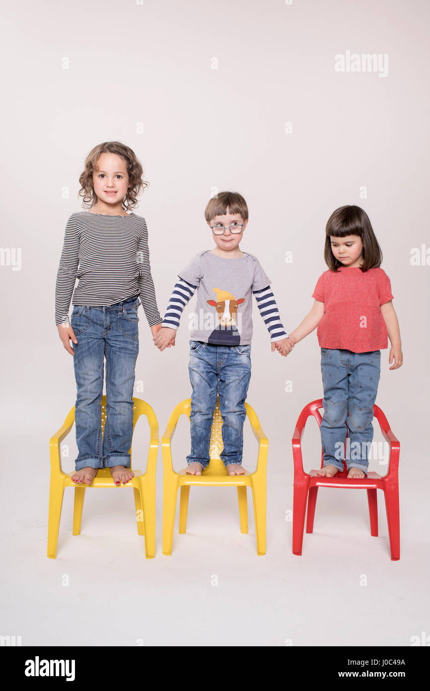 Ritratto di tre bambini in piedi sulle sedie colorate, tenendo le mani, studio shot Foto Stock