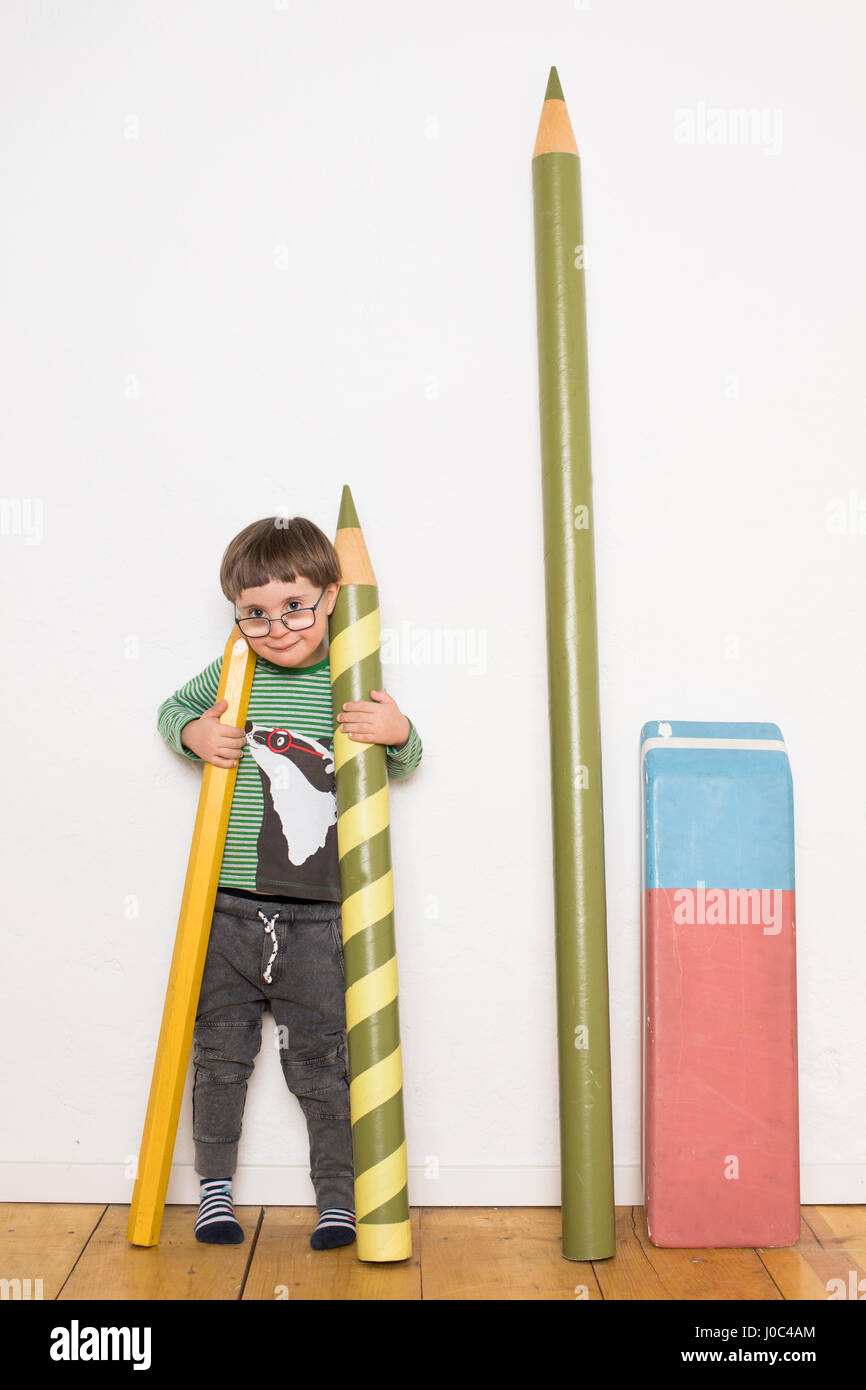 Giovane ragazzo in piedi, azienda di dimensioni giganti matite, cancelleria gigante appoggiata sulla parete accanto a lui Foto Stock