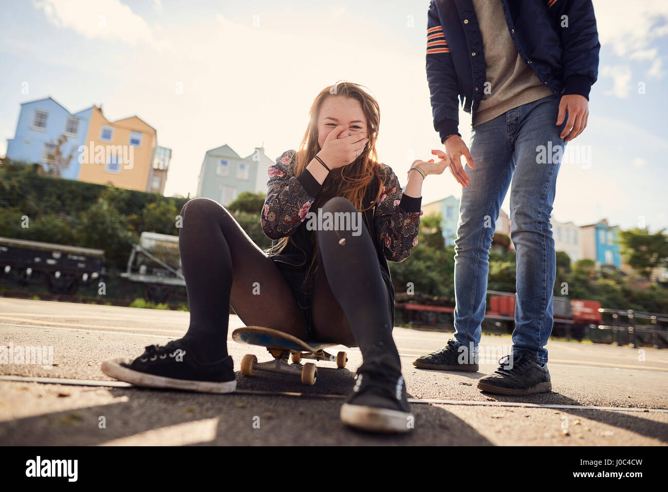 Due amici ingannare intorno all'aperto, giovane donna seduta su skateboard, ridendo, bassa sezione, Bristol, Regno Unito Foto Stock