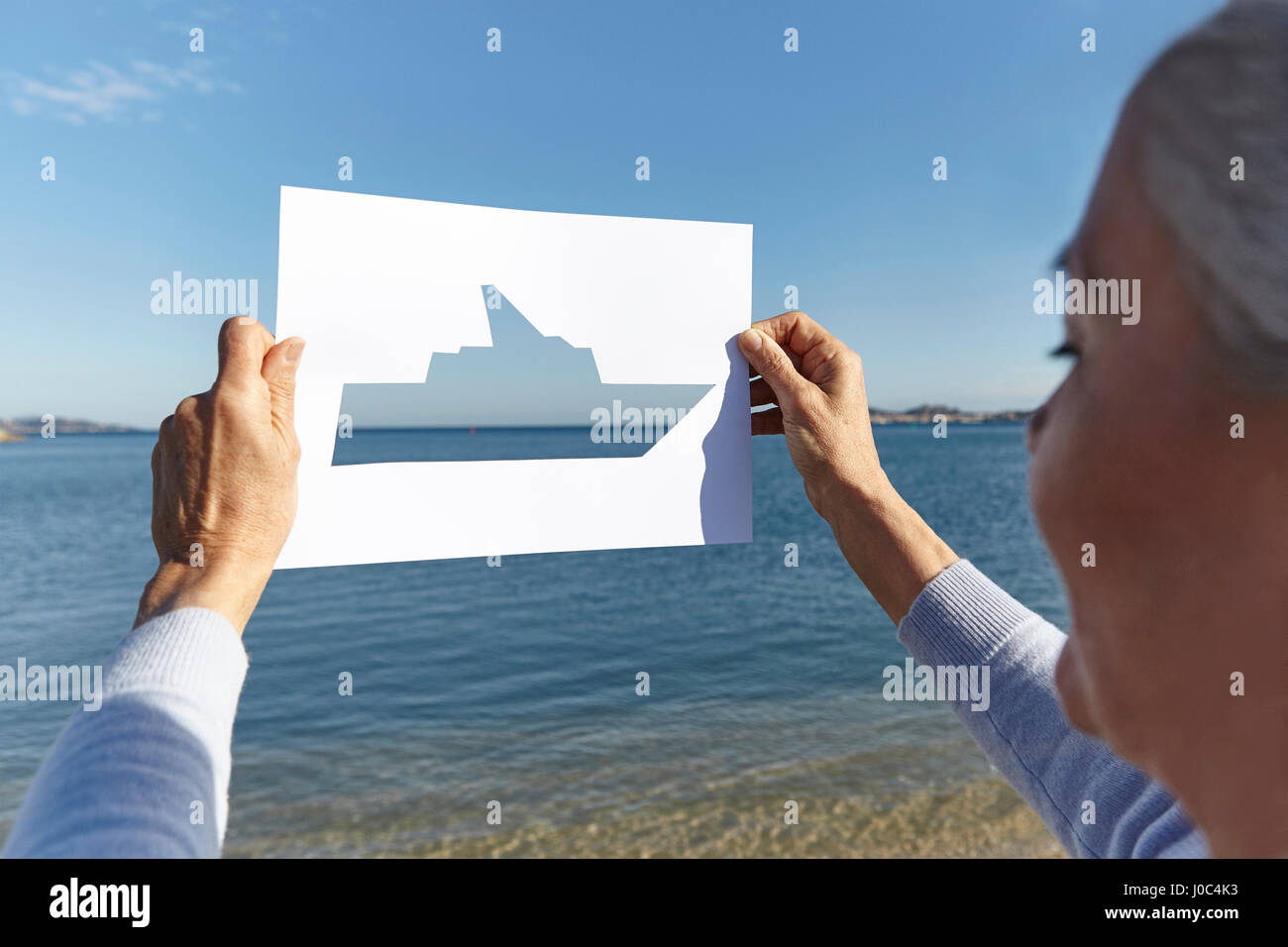 Senior donna in piedi di fronte al mare, tenendo fuori la carta con taglio-fuori della barca Foto Stock