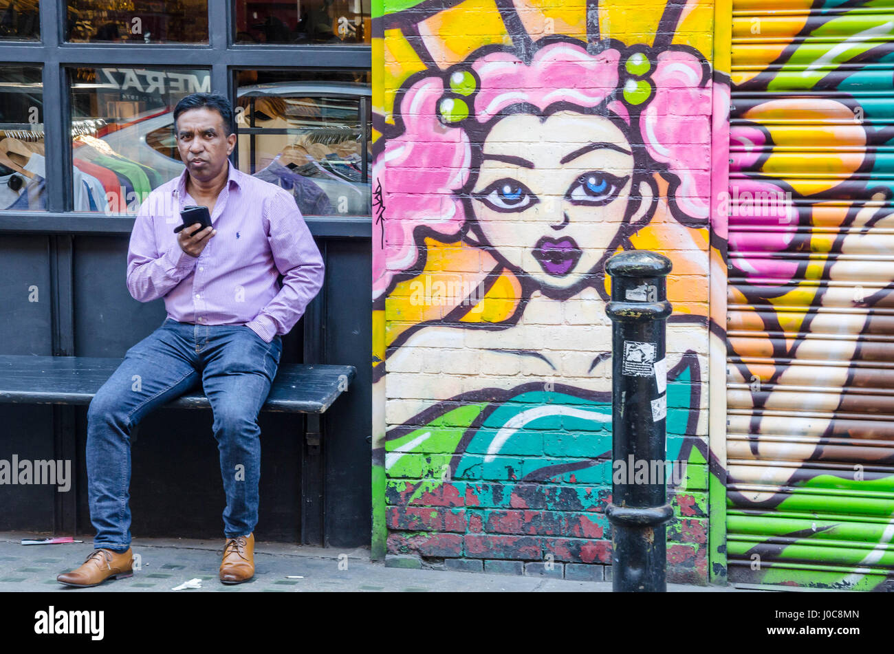 Un uomo si siede su una panchina accanto a un negozio di Hanbury Street nella zona est di Londra che è coperto in un luminoso murale colorato e controlla il suo telefono cellulare. Foto Stock