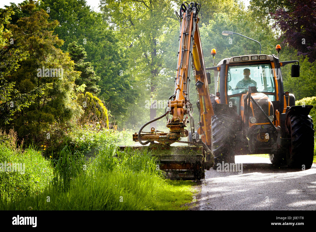 La falciatura di erba spallamento lungo la strada in uno spazio pubblico con grande arancione falciatrice trattore Foto Stock