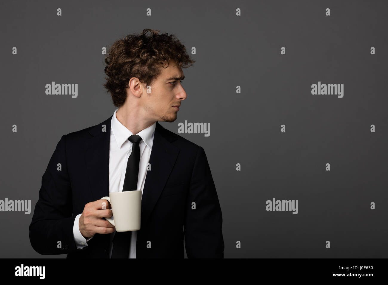Elegante parentesi di uomini in tuta tenendo una tazza di caffè o di tè in mani e guardando sopra la sua spalla, su sfondo grigio Foto Stock
