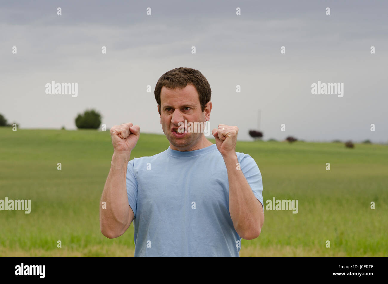 Angry Man all aperto con pugni serrata con un campo erboso dietro di lui. Foto Stock