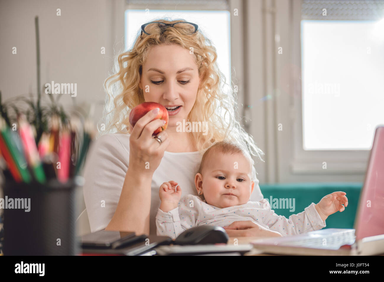 Madre mangiare red apple e utilizzando il portatile in ufficio a casa con il bimbo in braccio Foto Stock