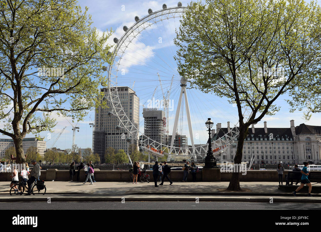 I turisti, corridori e altri pedoni passare lungo il Victoria Embankment al sole nel centro di Londra con il London Eye in background Foto Stock