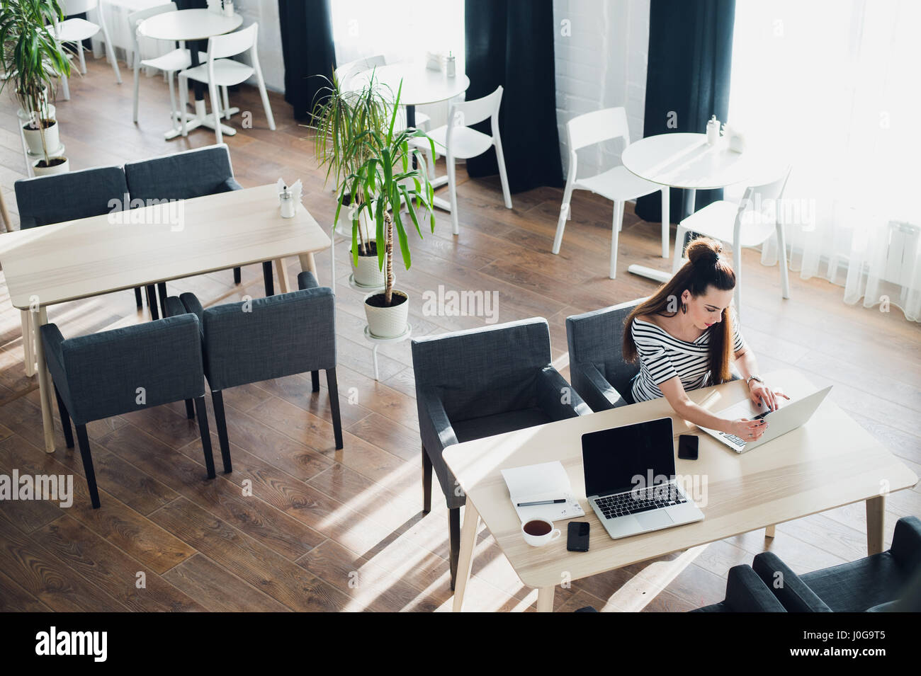 Tettuccio di colpo di giovane donna seduta al tavolo con una tazza di caffè e telefono cellulare navigando in rete sul suo computer portatile. Vista superiore della femmina usando il suo computer portatile a un cafe'. Foto Stock