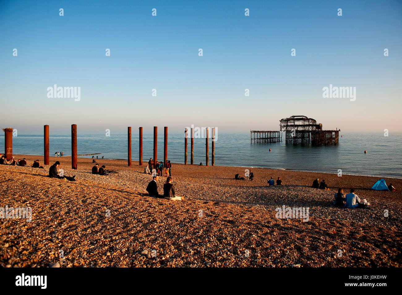 Persone sedersi sulla spiaggia di ciottoli a Brighton il Molo Ovest un famoso punto di riferimento sulla costa sud dell'Inghilterra, ora un bruciato arrugginimento scheletro. Foto Stock