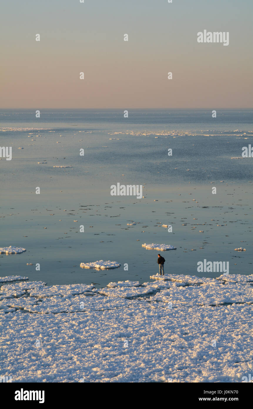 Un uomo si erge sul bordo di una lastra di ghiaccio e guarda giù verso l'acqua. La vastità del lago Michigan si estende al di là di lui. Foto Stock