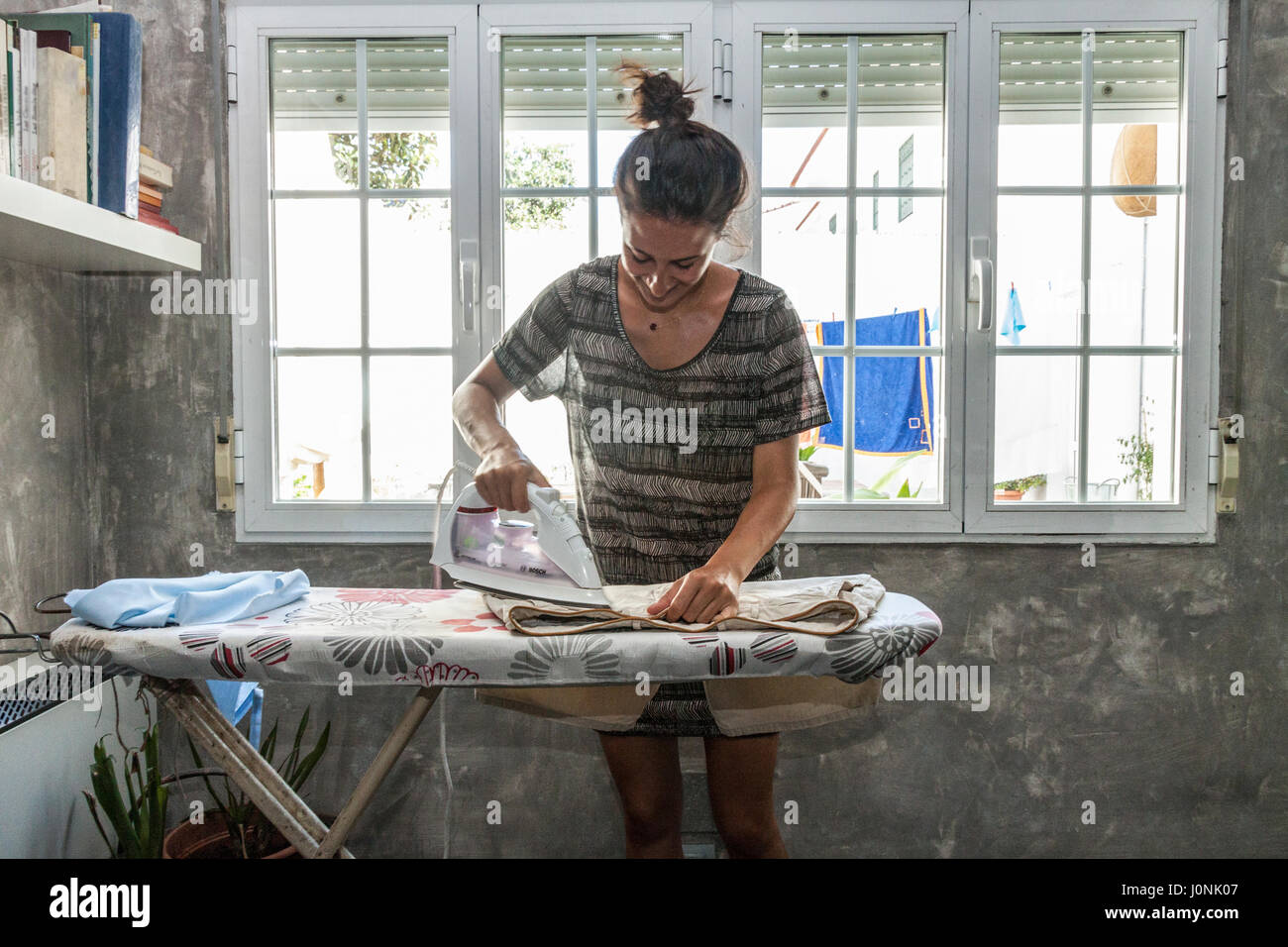 Home faccende. Una giovane e bella donna la stiratura di panni a casa, confortevolmente vestito, capelli legati in un panino. Foto Stock