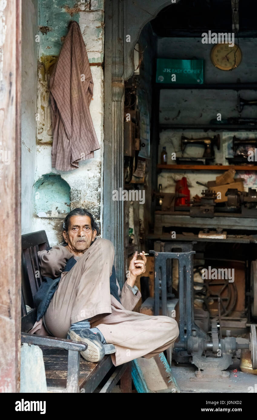 Negoziante fumatori, riposa in un negozio di riparazione per un vecchio stile vintage mano messo in moto le macchine da cucire, Pragpur heritage village, Himachal Pradesh, India Foto Stock