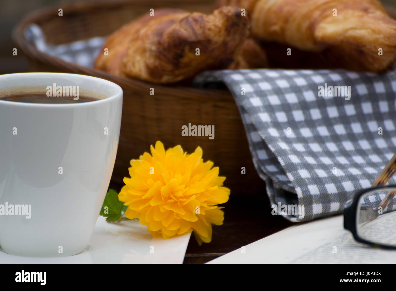 Close up di un cesto di croissant, una tazza di caffè con un piccolo fiore giallo accanto ad esso in un libro aperto e un paio di occhiali. Foto Stock