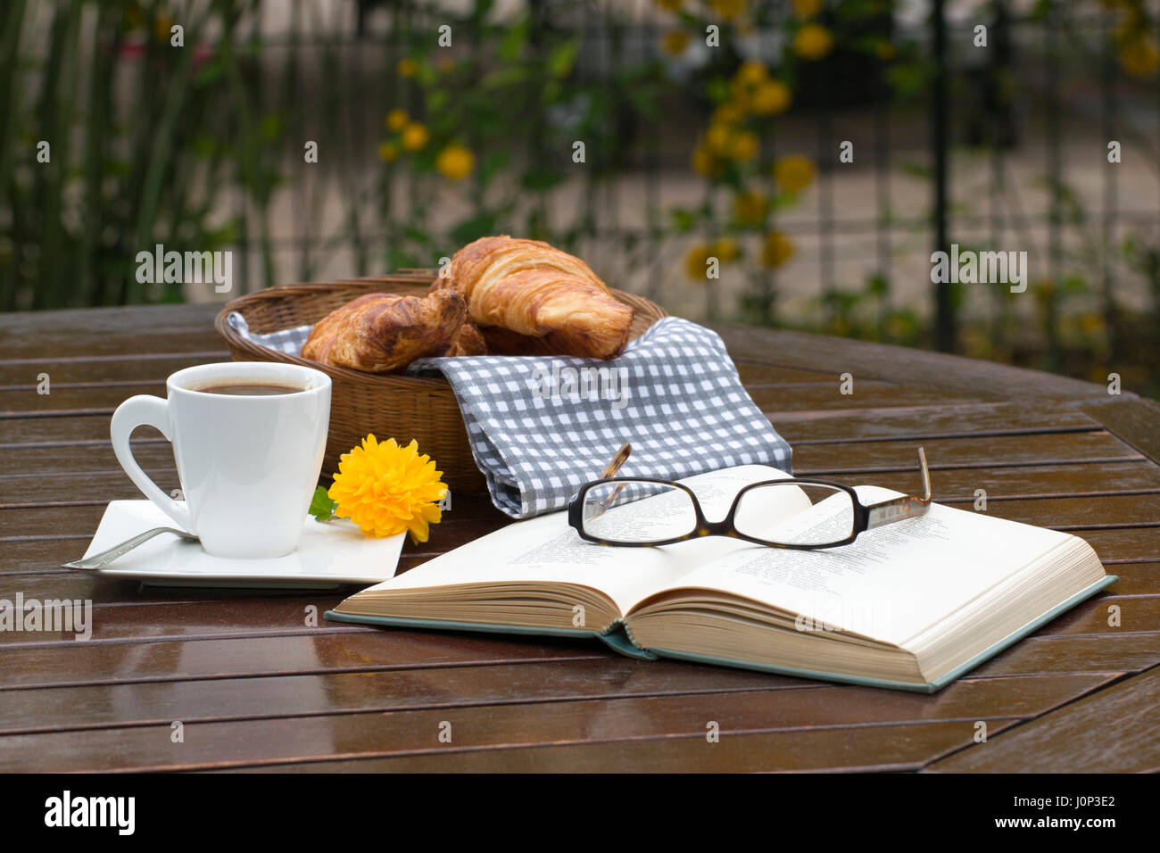 Un cesto di croissant, una tazza di caffè, un libro aperto e un paio di occhiali su una tavola di legno Foto Stock