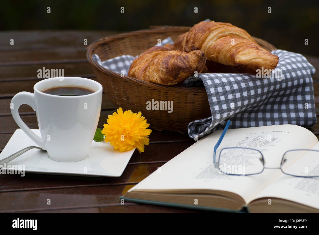 Un cesto di croissant, una tazza di caffè, un libro aperto e un paio di occhiali su una tavola di legno Foto Stock