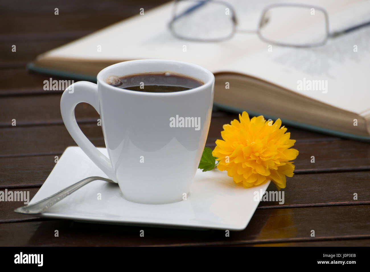 Una tazza di caffè con un giallo fiore piccolo e un cucchiaino Sul piattino, accanto a un libro aperto e occhiali su una tavola di legno, all'esterno. Foto Stock
