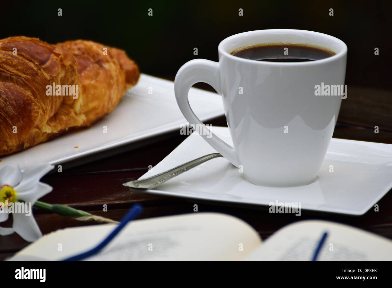 Una tazza di caffè nero e un croissant e un libro aperto con gli occhiali su di esso, un fiore su una tavola di legno, all'esterno. Foto Stock