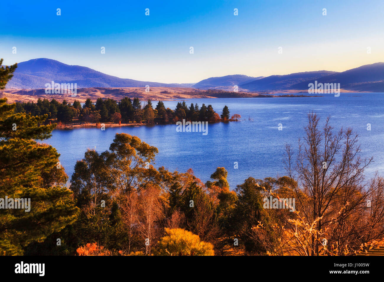 Acque blu del lago Jindabyne circondato da montagne innevate national park e luminose fronde di alberi in vista in elevazione a caldo la mattina dolci raggi del sole. Foto Stock