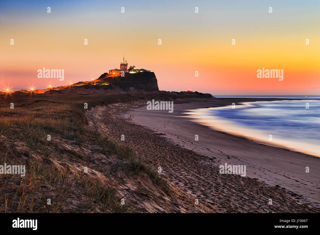 Dune di sabbia con erba a Nobbys beach, nobbys testa e nobbys faro in Newcastle, NSW, a colorato orange sunrise. Foto Stock