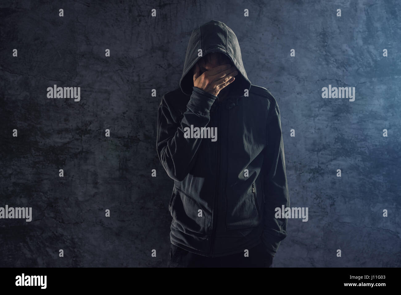 Premuto incappucciati giovani maschi adulti persona appoggiata sulla parete grungy e pianto Alone in the dark Foto Stock
