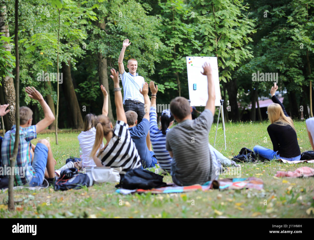 Un gruppo di giovani studenti nel parco. Vista di un uomo gesticulating con le sue mani, in piedi contro un gruppo di sfocati di persone sedute davanti a lui sull'erba. Foto Stock