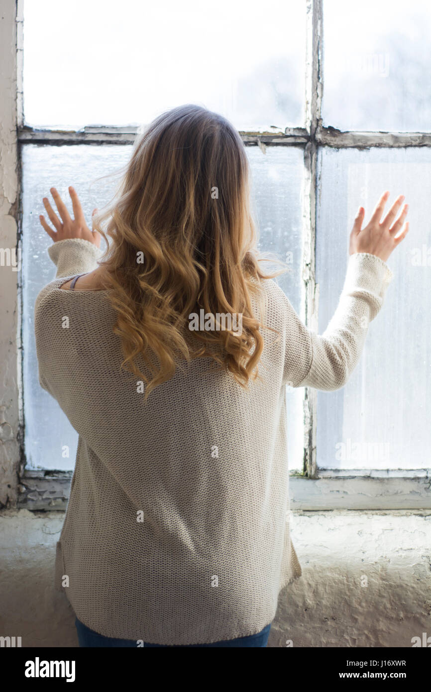 Vista posteriore di una donna bionda in piedi dalla finestra Foto Stock