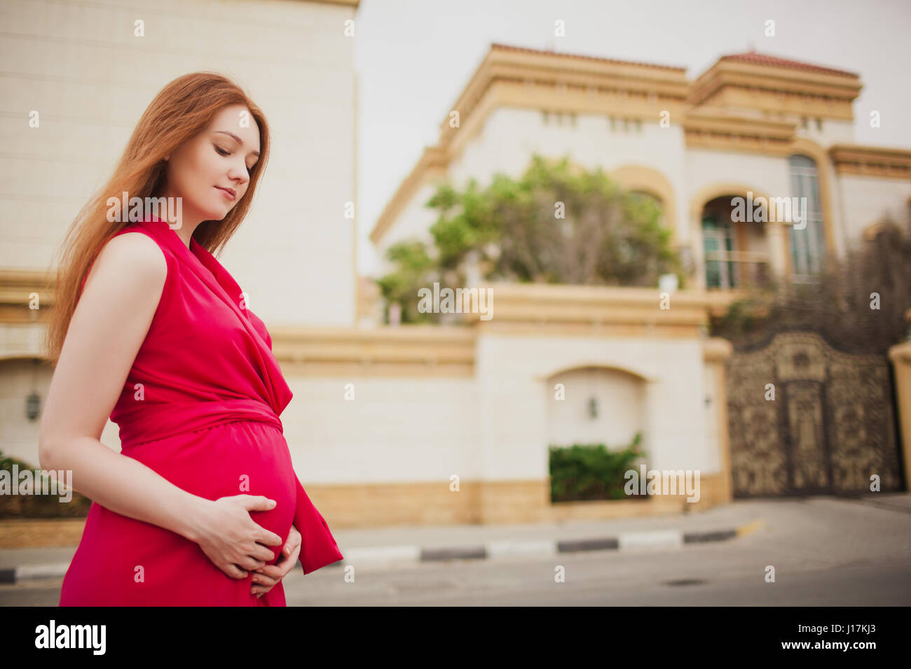 Carino i capelli rossi incinta ragazza in un abito rosso. Sullo sfondo della Villa. Foto Stock