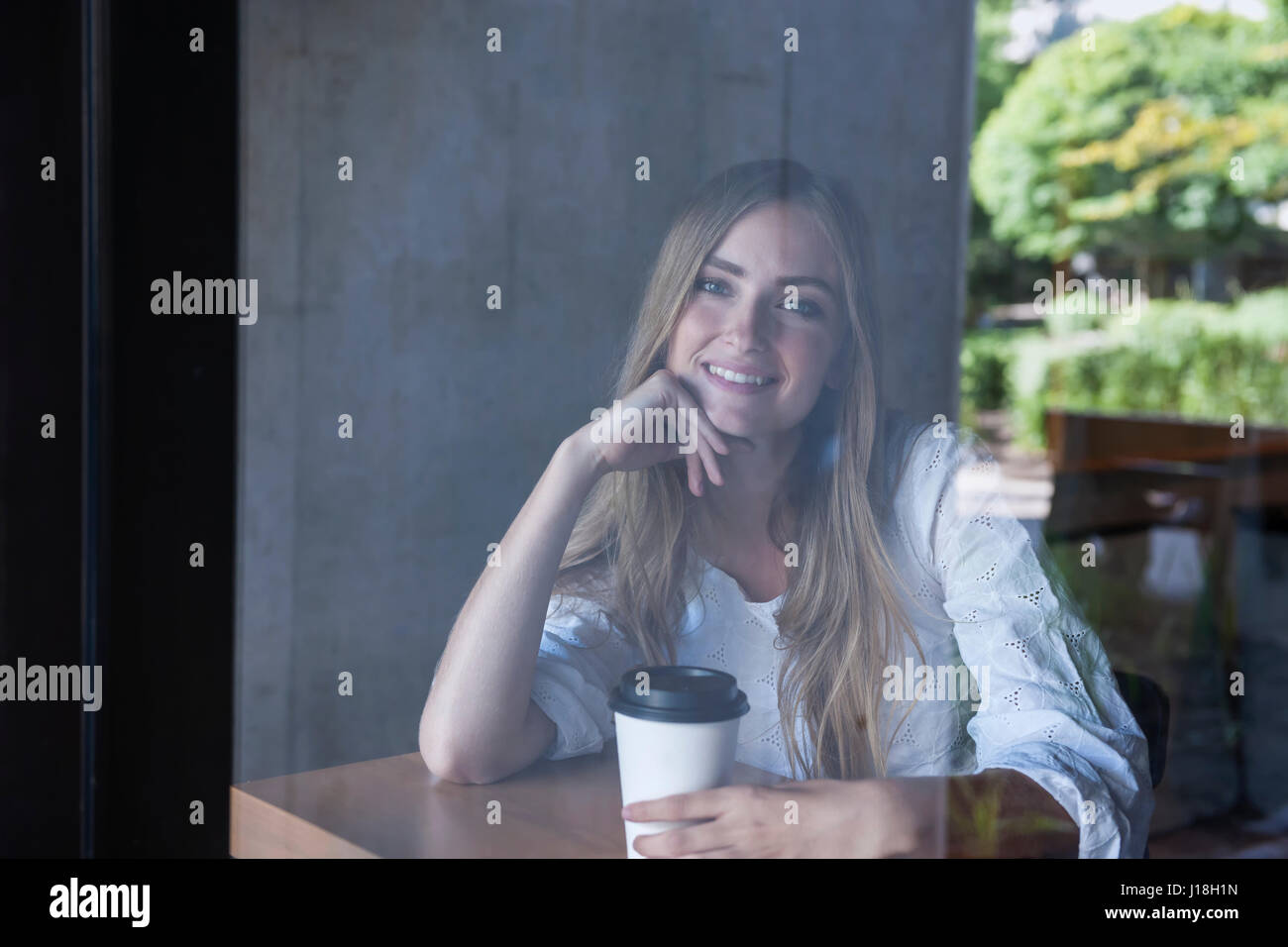Giovane donna sorridente seduto nella caffetteria con caffè fotografato attraverso la finestra di vetro da esterno Foto Stock