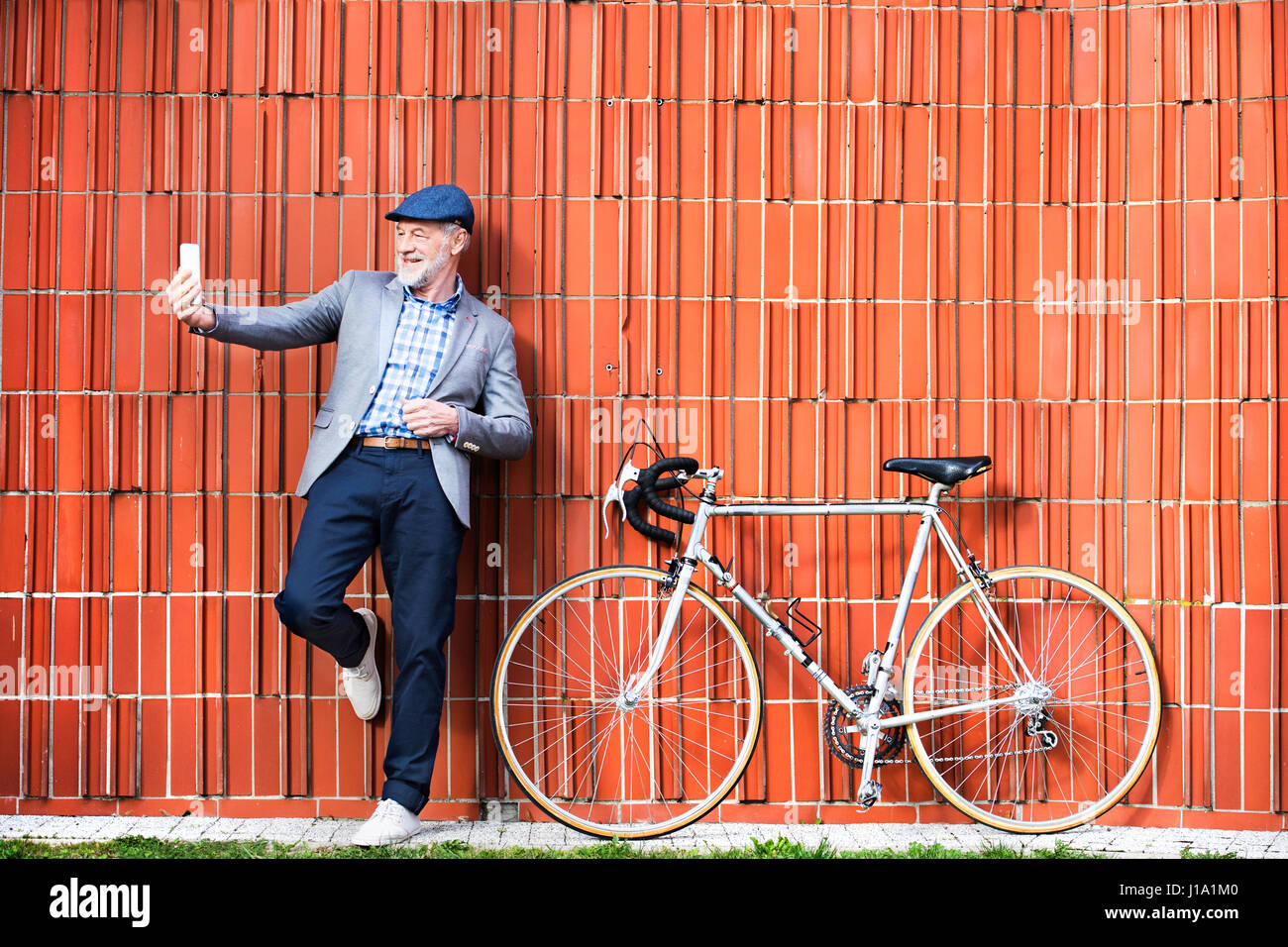 Bello il senior uomo in blu controllato camicia e giacca grigia con noleggio azienda smart phone, tenendo selfie. Orange muro di mattoni in background. Foto Stock