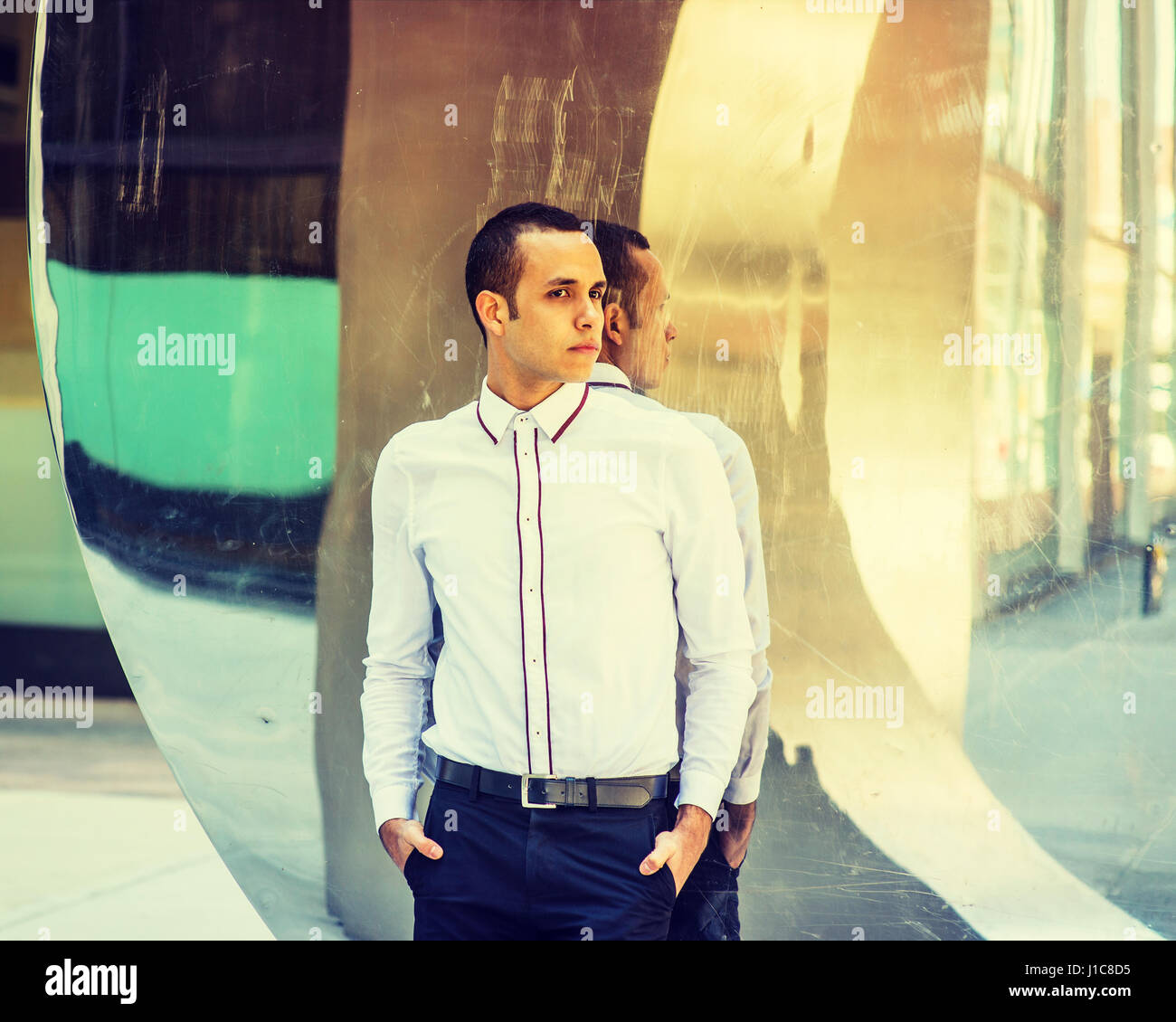 Giovane uomo americano che indossa una camicia bianca, pantaloni neri, due mani mettendo in tasche, in piedi contro il metallo specchio con riflessioni di New York, cercando Foto Stock