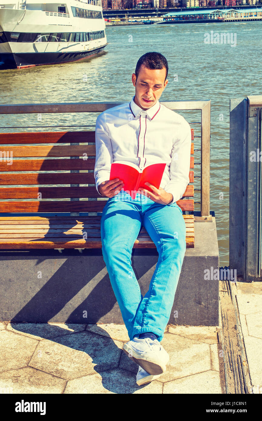 Giovane uomo americano in viaggio in New York, indossa una camicia bianca, pantaloni blu, bianco sneakers, seduti su una sedia sotto il sole dal fiume, lettura red book, RELA Foto Stock