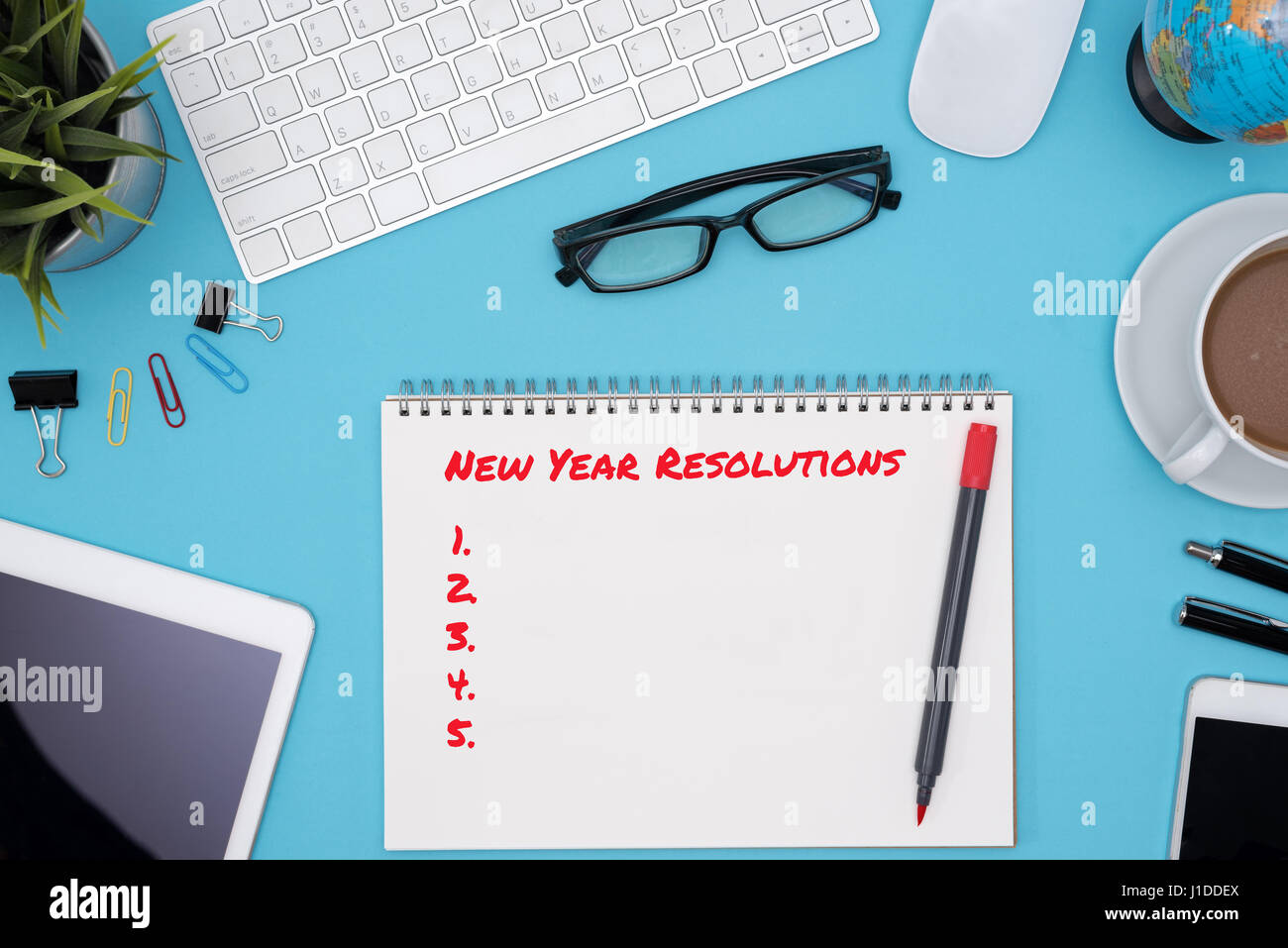 Propositi per l'anno nuovo concetto con spirale aperta notebook, computer, penne, telefono cellulare, tablet, fiore, occhiali, Globe e tazza di caffè sulla scrivania blu ta Foto Stock