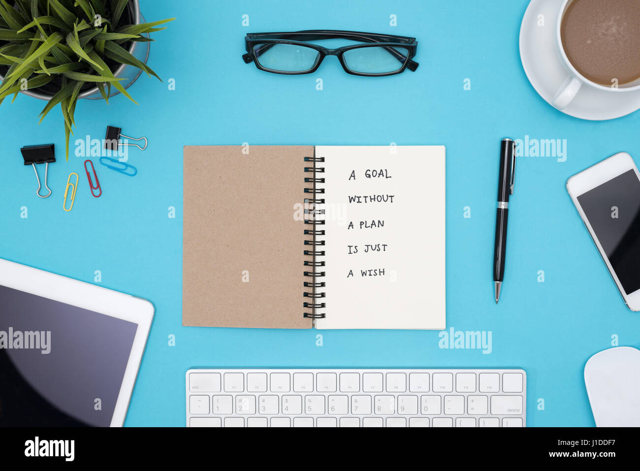 Un obiettivo senza un piano è solo un auspicio la grafia motivazionale sul notebook a spirale con computer, forniture per ufficio, occhiali e caffè sulla scrivania blu Foto Stock