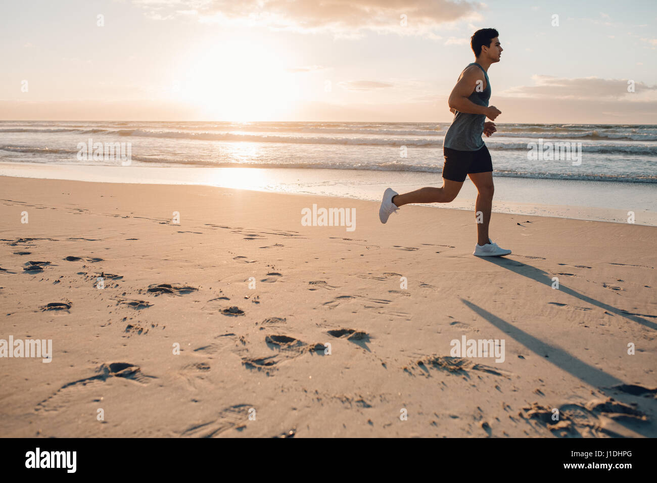 Vista laterale colpo di montare il giovane uomo che corre sulla spiaggia di mattina. Un sano modello maschile jogging sulla riva del mare. Foto Stock