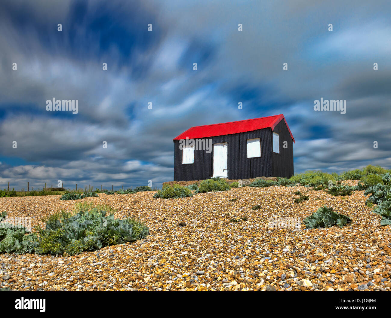 Un rosso brillante, il bianco e il nero shack su una spiaggia di ciottoli contro una lunga esposizione di cielo blu e nuvole. Foto Stock