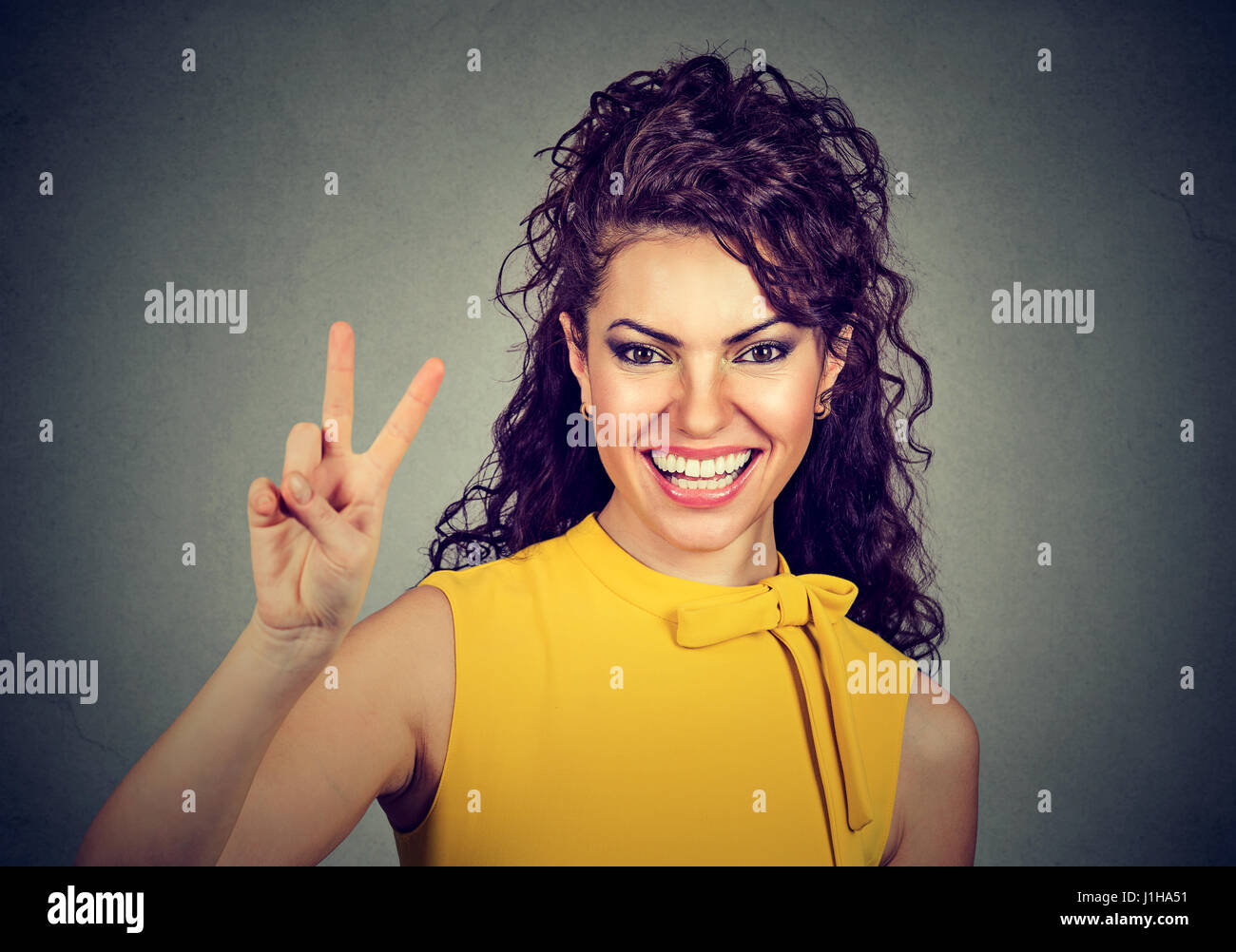 Donna sorridente in abito giallo che mostra la vittoria o segno di pace isolato sul muro grigio sfondo Foto Stock