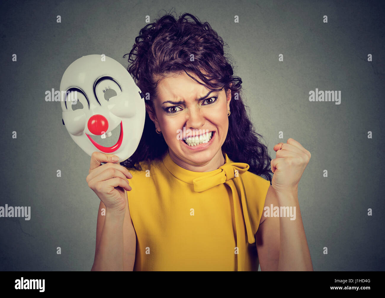 Ritratto arrabbiato urlando donna togliersi la maschera di clown esprimendo felicità isolato sul muro grigio Sfondo. Le emozioni umane sensazioni Foto Stock