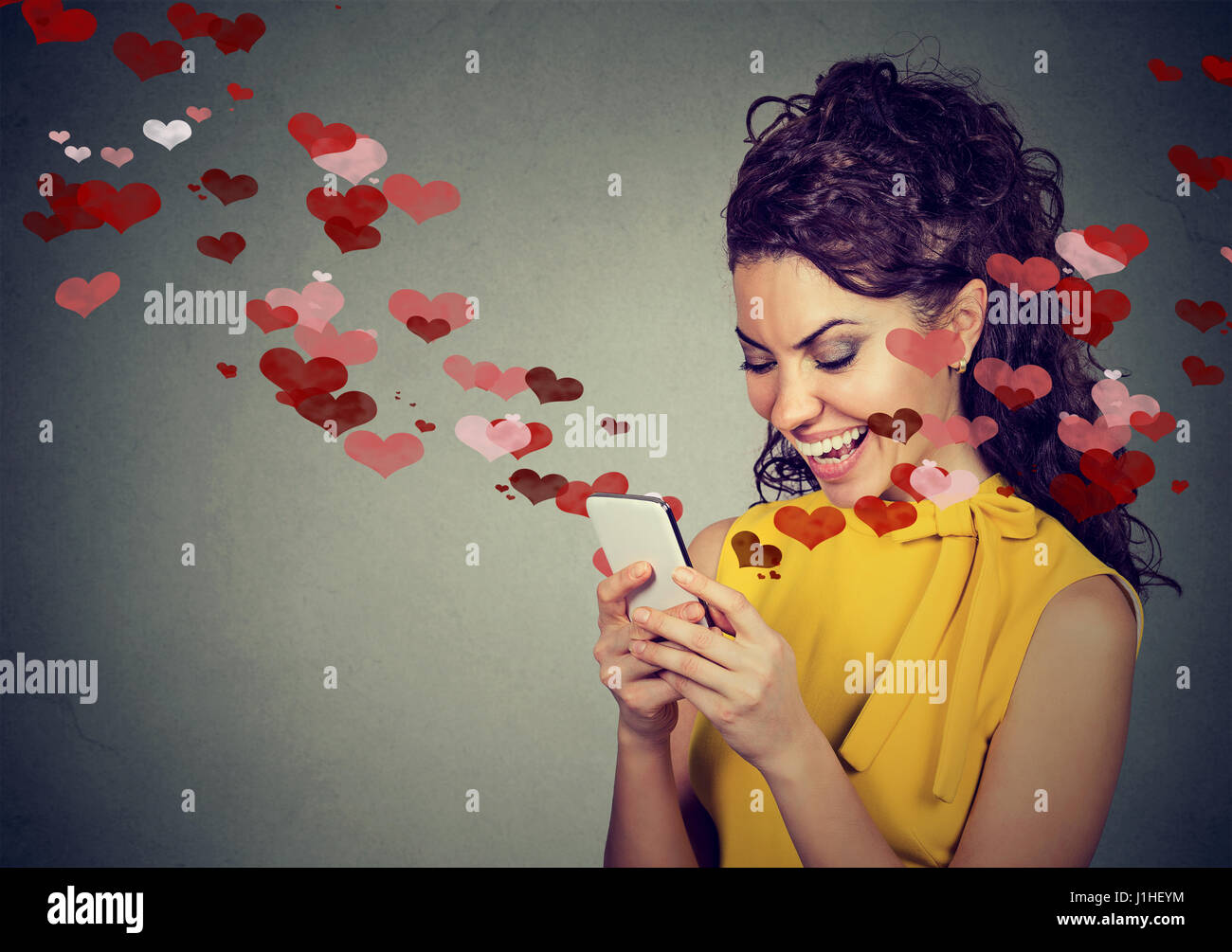 Bella donna felice amore invio messaggio di testo sul telefono cellulare con cuori rossi volare lontano dallo schermo isolato su sfondo grigio. Positivo e umano Foto Stock
