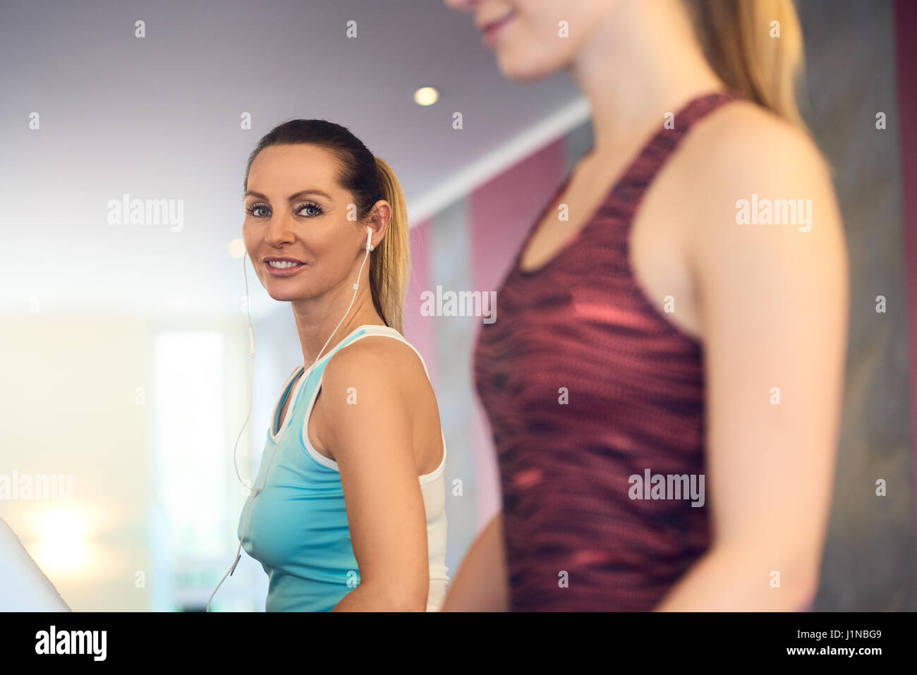 Attraente donna di mezza età con gli auricolari in piedi sul tapis roulant in palestra, guardando la fotocamera e sorridente con spazio di copia Foto Stock