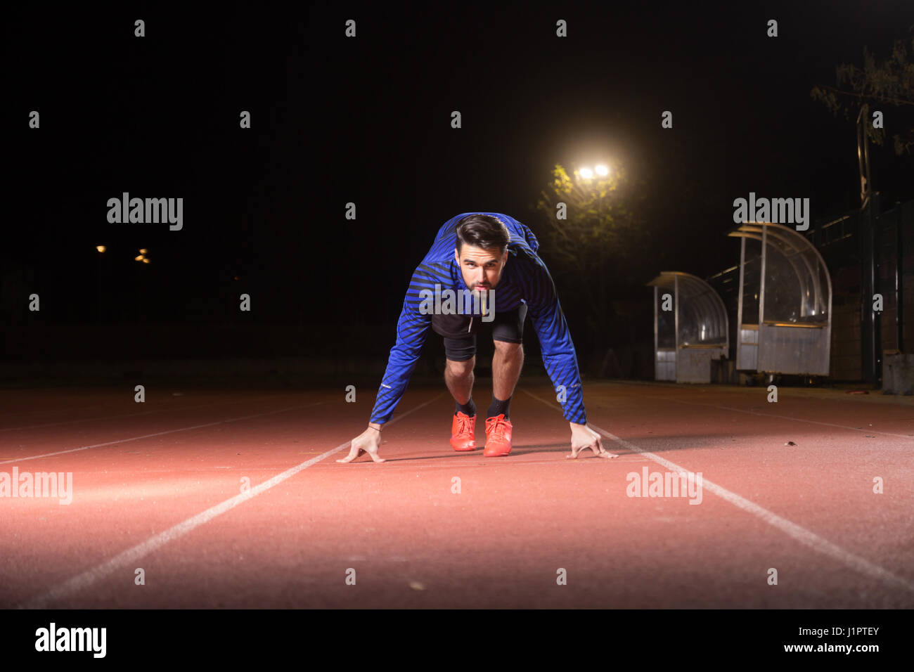 Un giovane uomo adulto, runner sprinter, posizione start, esecuzione di pista rossa. Tempo di notte scura, all'esterno. Foto Stock