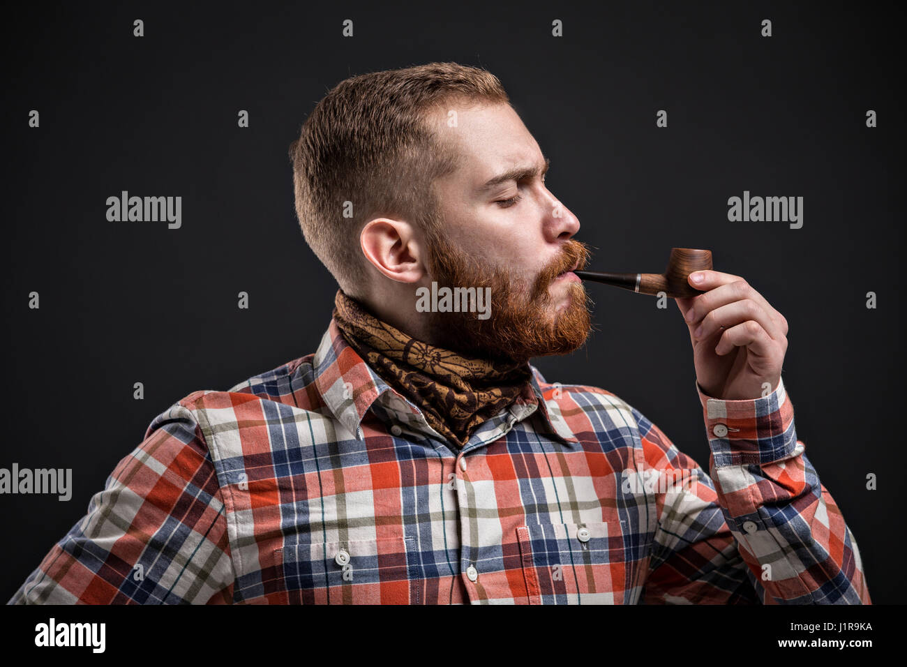 Adulto uomo barbuto in verifica shirt tubazione di fumare mentre guardando la fotocamera con un occhio Foto Stock
