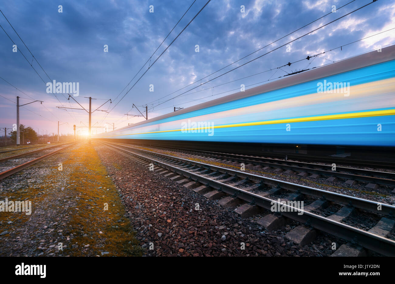 Alta velocità passeggeri blu treno in movimento su ferrovia via al tramonto. Sfocato il moderno treno. Stazione ferroviaria contro il drammatico cielo nuvoloso. Ferrovia tra Foto Stock