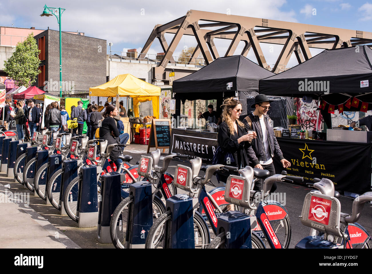 Street View di Brick Lane Market con hipster giovane e chioschi accanto a un cicli di Santander docking station. Brick Lane, Shoreditch, London, Regno Unito Foto Stock