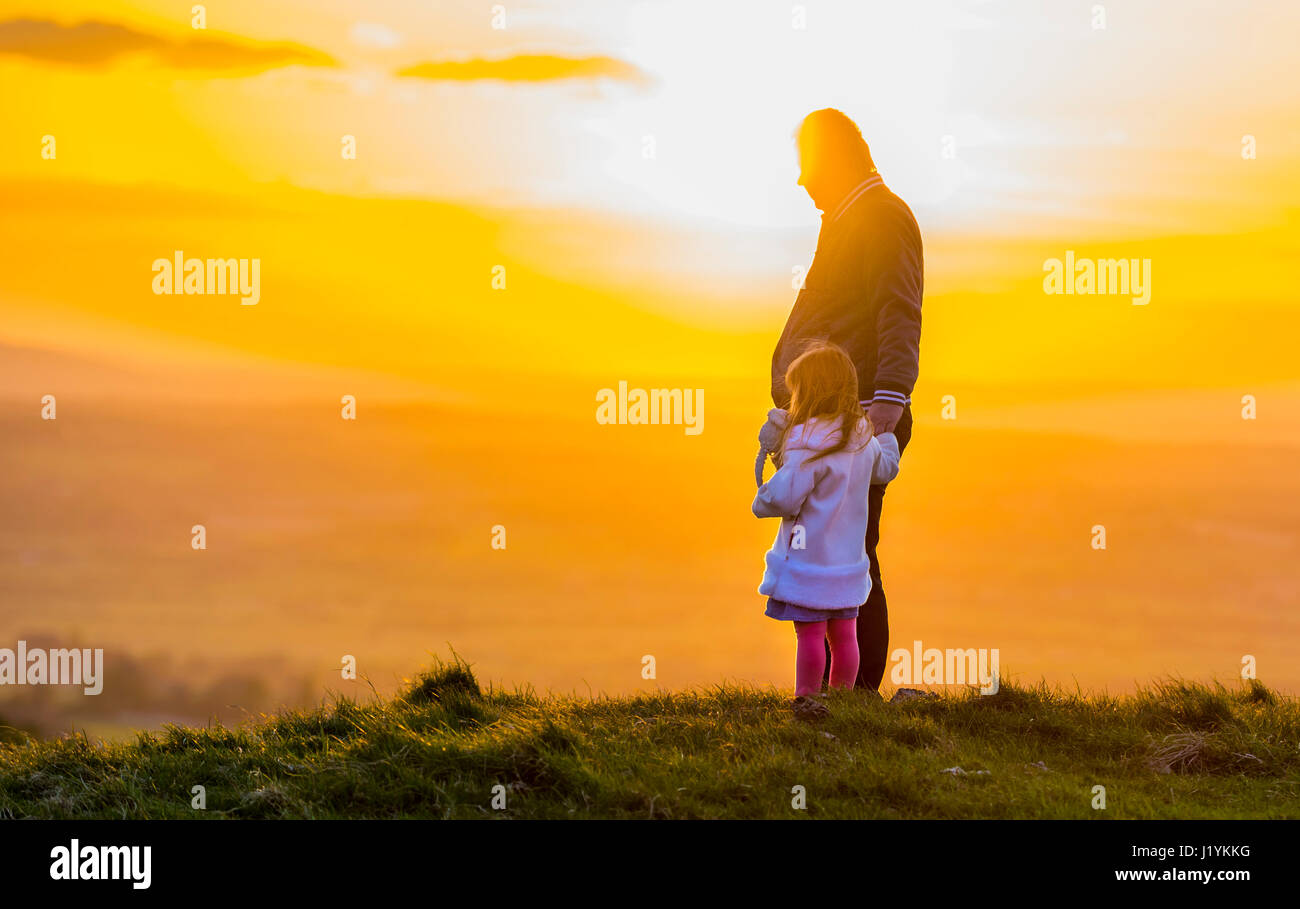 Single parent family. Padre e figlia insieme permanente tenendo le mani su una collina, godendo di guardare il sole andare giù. Foto Stock
