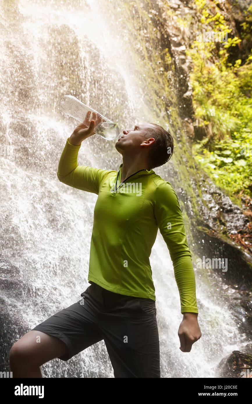 Escursionista uomo di bere acqua fresca pulita da un naturale. Giovane turista acqua potabile da una bottiglia cascata esterna in background. Bere acqua, hydr Foto Stock