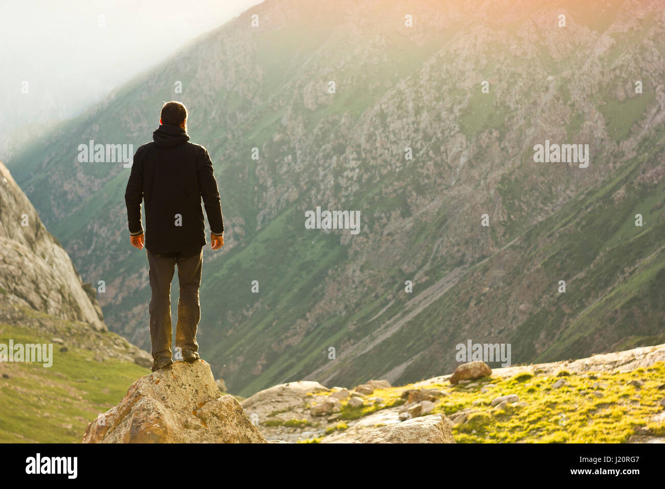 Uomo in camicia nera in piedi con le mani in alto al di sopra di pendii montani a giallo tramonto Foto Stock