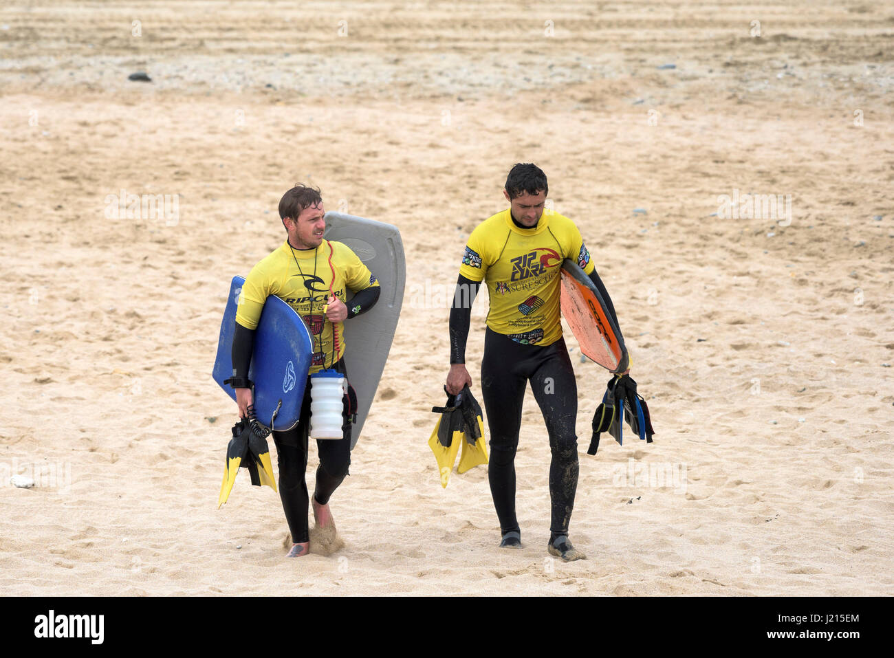 Due istruttori di surf a piedi in tutta la spiaggia Bodyboards scuola di surf di sabbia di attività per il tempo libero Fistral Cornwall Regno Unito Foto Stock