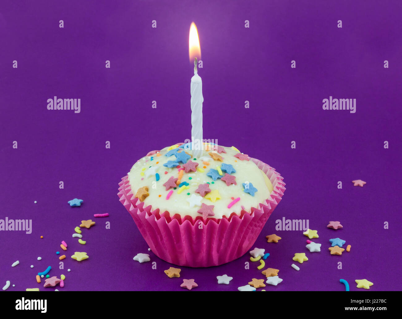 Viola luminoso con uno sfondo bianco singolo Cup cake in carta rosa caso e candela accesa con colore star spruzzino per la celebrazione di compleanno Foto Stock