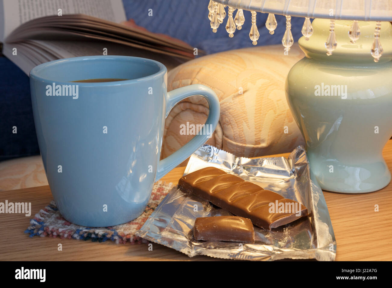 Lo stile di vita quotidiano hygge casalingo scena di una persona rilassarsi leggendo un libro con la tazza di caffè e cioccolato da una lampada da tavolo a casa. Inghilterra Regno Unito Gran Bretagna Foto Stock