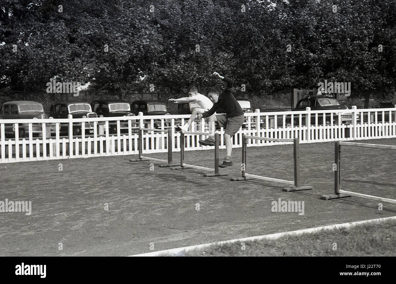 Anni '50, storico, fuori da una pista da corsa di cinder, due giovani ragazzi che gareggiano in una corsa di ostacoli saltano sopra barriere o ostacoli, con automobili dell'epoca parcheggiate in una linea dietro una recinzione bianca, Inghilterra, Regno Unito. Foto Stock