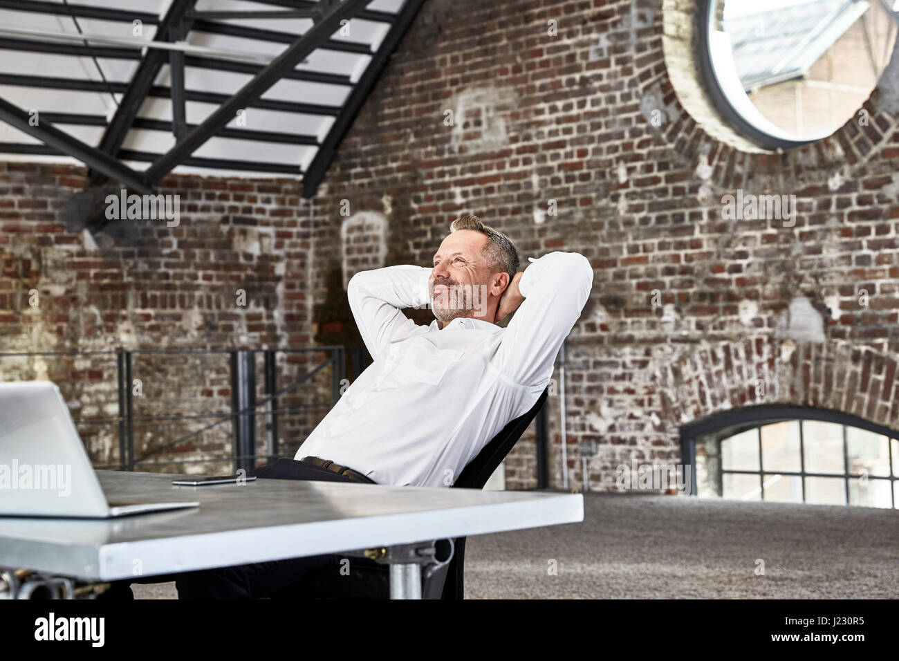 Imprenditore sorridente seduta a tavola in un loft Pendente ritornare Foto Stock